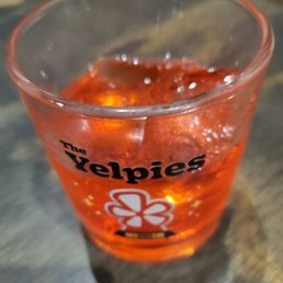 A clear glass containing a red-colored drink with ice cubes, labeled 'The Helpies,' placed on a dark, textured surface.