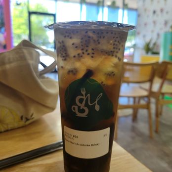 Sip Munch Cafe - Artichoke water - A large plastic cup containing an artichoke-flavored beverage with tapioca pearls, placed on a wooden table inside a cafe with bright windows. - Coffee & Tea Near Me - Austin, Texas