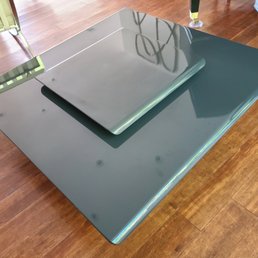 A glass table with a smaller glass cover on top is situated on a wooden floor, with chairs and other furniture visible nearby.
