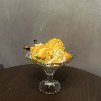 Mango Mango Dessert - Mango Triple Sundae - A mango-flavored ice cream sundae topped with whipped cream, caramel drizzle, and chocolate wafer rolls, served in a glass dish on a dark wooden table. - Desserts Near Me - Edison, New Jersey