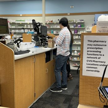 CVS Pharmacy - Takes two people to stare at a computer while one works. - A person in a plaid shirt and jeans is at the CVS pharmacy checkout, working on the register, while another person stands behind them. - Drugstores Near Me - Gainesville, Virginia