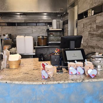 Rice Box & Boba  - A cafe counter with a cash register, snack packages, and decorative piggy banks, set against a kitchen backdrop with kitchenware and containers. - Laotian Near Me - Albuquerque, New Mexico
