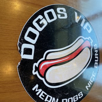 Dogos VIP - Eat here - A round sticker with a black background displays a stylized hot dog graphic with toppings and the text 'Dogos VIP' at the top. Additional text around the edge reads 'Medn Dogs Nice BunS.' The sticker appears worn with visible scratches and is placed on a wooden surface, representing branding for a hot dog and birria-focused eatery in Albuquerque, NM. - Hot Dogs Near Me - Albuquerque, New Mexico