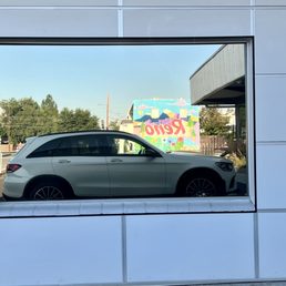 Before - The image shows a silver SUV parked outside, reflected in a large, rectangular window of a modern building with white paneling. In the background, a colorful mural with the word 'Revo' and various shapes and trees can be seen, along with a clear blue sky and some utility poles.