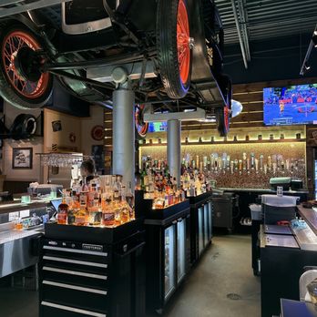 Ford's Garage Florence - Bar - A vintage black car with red wheels is mounted upside down on the ceiling of a bar, with a bar counter and liquor bottles below. - Burgers Near Me - Florence, Kentucky