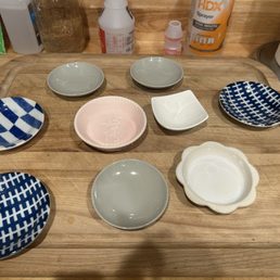 Various sizes of dishes for feeding my foster kittens - A collection of small ceramic dishes with various shapes, colors, and patterns is displayed on a wooden tray. The dishes include checkered blue and white patterns, plain gray, a pink dish with a script, and a white scalloped-edged dish. The background shows kitchen items such as a paper towel roll, spray bottle, and other containers.