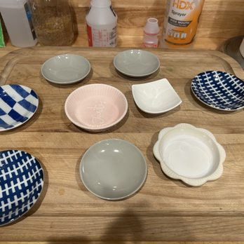 Daiso - Various sizes of dishes for feeding my foster kittens - A collection of small ceramic dishes with various shapes, colors, and patterns is displayed on a wooden tray. The dishes include checkered blue and white patterns, plain gray, a pink dish with a script, and a white scalloped-edged dish. The background shows kitchen items such as a paper towel roll, spray bottle, and other containers. - Department Stores Near Me - Concord, California