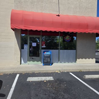 Mandarin House - The image shows the exterior of Mandarin House, a restaurant with a beige stucco facade and a large red fabric awning. The glass entrance door has a small sign and a Pepsi advertisement board outside, promoting an all-day buffet priced at $17.99. The parking lot in front has marked parking spaces and a concrete wheel stop. The reflection in the glass shows trees and a person taking the photo. The sky is clear and blue. - Chinese Near Me - Sonora, California