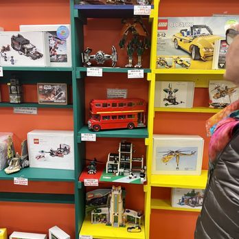 Brick It Again - Display of a few builds, and boxed Lego sets - Multiple brightly colored shelves showcase an assortment of LEGO sets and models, including themed sets, vehicles, and custom builds. The display includes boxed LEGO products, assembled models, and promotional signs, with some items marked as sold. The store features a vibrant, organized layout with orange, green, and yellow shelving units, and a partial view of a customer on the right side of the image. - Toy Stores Near Me - Valley Cottage, New York
