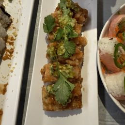 A rectangular white plate holds a sushi roll topped with diced avocado, cilantro leaves, and a drizzle of sauce. The roll appears to be fried or tempura-style, with a crispy exterior. Part of another sushi dish is visible on the left, and a bowl with rice and vegetables is partially seen on the right. The setting suggests a restaurant serving Japanese or Asian cuisine.