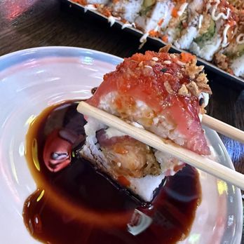 Dragon Sushi - Amazing roll piece - A piece of sushi is held with chopsticks over a small dish of soy sauce. The sushi features rice, fish, and toppings, with a drizzle of sauce and garnishes. In the background, a large platter of assorted sushi rolls is visible on a dark wooden table. - Sushi Bars Near Me - Albuquerque, New Mexico