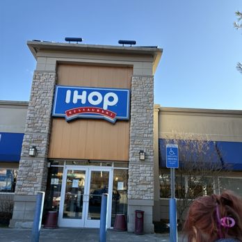 IHOP - Outside - The photo captures the exterior of an IHOP restaurant building with a prominent blue and white sign displaying the IHOP logo and the word 'RESTAURANT.' The building features stone accents around the entrance, glass doors, and a handicap parking sign nearby. The sky is clear and blue, and part of a person's head with reddish hair and a hair clip is visible in the lower right corner. - Breakfast & Brunch Near Me - Orange, Connecticut