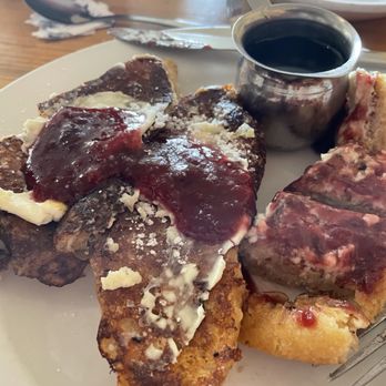 Tune-Up Cafe - French toast - A plate features French toast topped with berry jam, butter, and powdered sugar. Next to the French toast are slices of pizza with cheese and tomato sauce. A small metal cup contains black coffee. The setting appears to be a casual dining environment with a wooden table and utensils nearby. - New Mexican Cuisine Near Me - Santa Fe, New Mexico