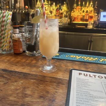 Fulton Hall - Piñata - A tropical cocktail with a lemon wedge and striped straw is placed on a bar counter, with bottles and bar equipment in the background. - Pubs Near Me - Brooklyn, New York