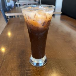 A tall, transparent glass contains iced coffee with ice cubes, placed on a wooden table. The background shows a modern restaurant interior with tables, chairs, and a neutral color scheme.