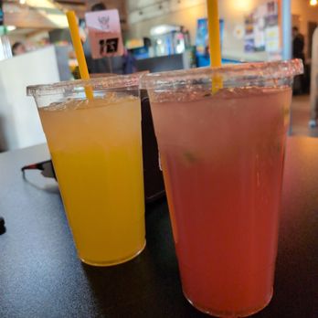 Adis - Mango lemonade & Strawberry watermelon - Two large cups of fruit beverages are placed on a dark table inside a restaurant or cafe. The left cup contains a yellow drink, and the right cup contains a pink drink. Both cups have clear plastic lids and yellow straws. The background shows a lively indoor setting with blurred figures, colorful lighting, and wall decor, indicating a casual dining environment. - Ice Cream & Frozen Yogurt Near Me - Tucson, Arizona