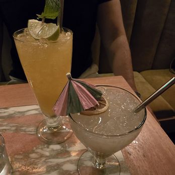 Daydream - The image shows two cocktails on a wooden table in a dimly lit setting. The tall drink is yellowish, garnished with lime wedges, mint, and a straw. The shorter drink is clear, with ice, a dried citrus slice, and a small paper umbrella. A person wearing a black shirt is seated behind the drinks, partially visible. The background includes a cushioned seat and a curtain, suggesting an indoor bar or lounge environment. - Cocktail Bars Near Me - Albuquerque, New Mexico