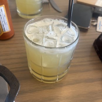 The Harvest Room - Booty Collins - A clear glass filled with iced lemonade, ice cubes floating on top, and two black straws, placed on a wooden table at a restaurant. - New American Near Me - Munster, Indiana