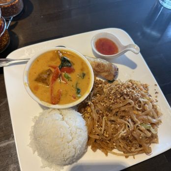 Dillon Thai restaurant - lunch combo with panang curry - A Thai cuisine meal served on a white square plate includes a bowl of yellow curry with vegetables and possibly chicken, a portion of steamed white rice, stir-fried noodles topped with crushed peanuts, and a spring roll accompanied by a small dish of sweet chili sauce. The presentation highlights typical Thai dishes with vibrant flavors and ingredients. - Thai Near Me - Dillon, Colorado