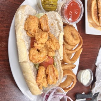 Cormier’s Kitchen - A seafood po'boy with fried shrimp, onion rings, pickles, and condiments on a white plate at a restaurant. - Southern Near Me - Spring, Texas