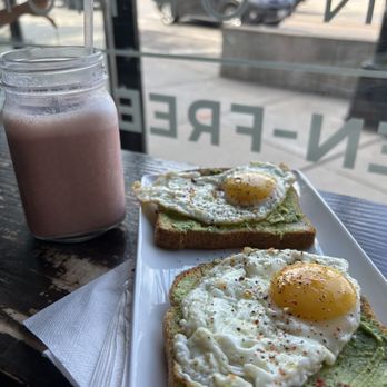 Oromo Cafe - Pistachio Strawberry - The image shows a cafe setting with two slices of avocado toast topped with fried eggs on a white rectangular plate. A mason jar filled with a pink smoothie and a straw is placed beside the plate. The table is near a window, revealing parked cars and a sidewalk outside. The window has partial lettering visible, indicating the cafe's interior view. - Coffee & Tea Near Me - Chicago, Illinois