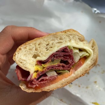 Calisto's - pastrami sandwich - A sandwich with sliced meats, cheese, lettuce, tomato, and pickles is held in a person's hand, with a paper bag and a table in the background. - Delis Near Me - Boston, Massachusetts