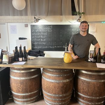 Springhill Cellars - I highly recommend having a designated driver most fun I had in ages - A man with short hair and a beard stands behind a bar made of three large wooden wine barrels, with a wooden countertop on top. The bar is situated in an indoor setting with a blackboard menu listing wines and prices behind him. The counter has several wine bottles and a yellow pitcher. The background includes a white wall, a curtain, and some hanging paper lanterns. The setting appears to be a wine tasting or retail space at Spring Hill Cellars in North Albany, Oregon. - Wineries Near Me - Albany, Oregon