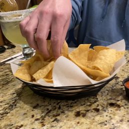 Chips and salsa! - A hand is seen grabbing tortilla chips from a basket lined with paper at a restaurant, with salsa and a drink on the table.