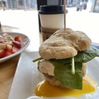 Cafe 301 - Fried Green Tomato Sandwich. - A fried green tomato sandwich features a crispy fried green tomato, fresh leafy greens, and a runny egg on a white rectangular plate. The sandwich is held together with a wooden skewer. In the background, there is a disposable coffee cup with a black lid and a bowl of strawberries with cream. The setting appears to be a cafe with large windows allowing natural light. - Food Trucks Near Me - Mountjoy, Pennsylvania