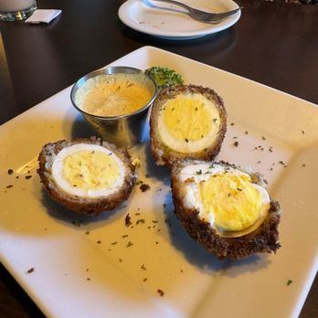 Dublin's Pass - Scotch Egg appetizer - A white square plate holds three Scotch eggs, each with a crispy breaded exterior and a soft-boiled egg inside. The eggs are sliced in half, revealing the yolk and white. A small metal container of mustard sauce is placed on the plate, garnished with a sprig of parsley. The setting appears to be in a restaurant, with a person sitting at the table and an empty plate with a fork in the background. - Irish Near Me - Springfield, Missouri
