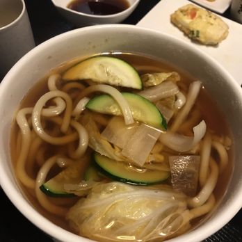 Araya Sushi - Plain Udon ($19.99 incl. dynamite roll) - A bowl of udon noodle soup contains thick noodles, zucchini slices, cabbage, and onions in a broth, with side dishes and dipping sauce visible. - Sushi Bars Near Me - Markham, Ontario