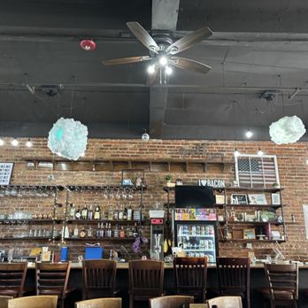 Loading Gastrobrunch - The image shows the interior of a restaurant with an industrial style, including exposed brick walls and a dark ceiling with a ceiling fan. Hanging light bulbs and cloud-shaped decorations are suspended from the ceiling. The bar area has bottles of alcohol, glasses, and a small refrigerator. There are wooden chairs lined up at the bar, and some tables and chairs are visible in the foreground. The decor includes a framed American flag and a sign that says 'I ❤️ BACON.' - Breakfast & Brunch Near Me - St Cloud, Florida
