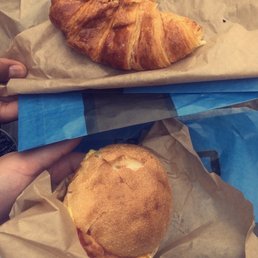 This was my brunch sandwich and my aunt was a ham and cheese on a croissant - The image shows two hands holding different food items wrapped in paper. The top hand holds a golden-brown croissant on brown parchment paper. The bottom hand holds a sandwich with a round, toasted bun, wrapped in brown paper. The background includes blue and brown paper, suggesting a casual food setting.