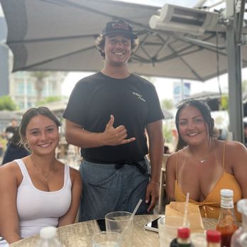 High/Low - Jacob was our server! - Two women are seated at an outdoor restaurant table, smiling at the camera. The woman on the left is wearing a white tank top, and the woman on the right is wearing a yellow spaghetti strap top. A man is standing behind them, smiling and making a shaka sign with his right hand. The table has various condiments, a smartphone, and drinks. The background shows other diners, umbrellas, and a cityscape with buildings and palm trees. - Breakfast & Brunch Near Me - Oceanside, California