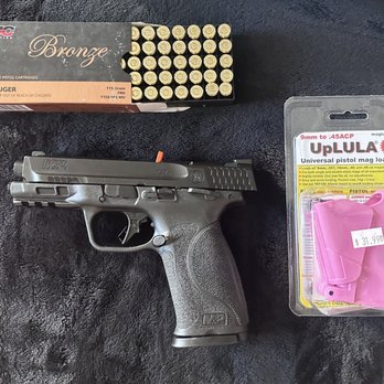 Guns Direct - First Hand Gun - A semi-automatic handgun with a textured grip and slide is positioned on a black surface, alongside a box of 9mm ammunition and a pink pistol magazine loader in its packaging. The ammunition box contains 50 rounds of 9mm Luger cartridges, and the magazine loader is priced at $31.99. The items are arranged neatly, with the ammunition box at the top, the handgun in the center, and the magazine loader on the right. - Outdoor Gear Near Me - Burbank, California