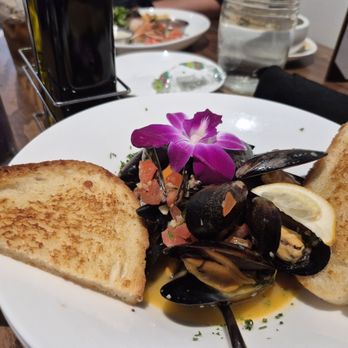 Harrison’s - Mussels - A seafood dish with cooked mussels served on a white plate, garnished with a purple edible flower and a lemon wedge. Two slices of toasted bread are placed on either side of the plate. The dish appears to be part of a meal at a restaurant, with other plates and drinks visible in the background. - Steakhouses Near Me - Indianapolis, Indiana