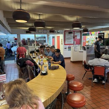 Duran Central Pharmacy - The image shows an indoor dining area with customers seated at a curved wooden counter and at tables. Several people are eating or waiting, with some wearing sunglasses and casual clothing. The decor includes hanging black pendant lights, tiled flooring, and a visible kitchen or service area in the background. The setting appears to be a casual restaurant or cafe. - Mexican Near Me - Albuquerque, New Mexico