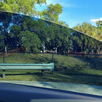 Jonathan Window Tint - Perfect edges! - A car window with dark tinting reflecting trees, sky, and clouds, set against a background of greenery and a fence. - Home Window Tinting Near Me - Boca Raton, Florida