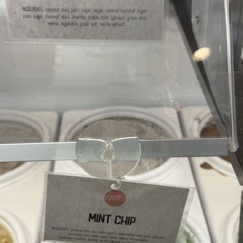 Gati - A sign with the label 'Mint Chip' is attached to a glass display case at Gati, an ice cream shop. The sign lists ingredients such as coconut milk, organic avocado, and chocolate chip, and is secured with a clear adhesive. The background shows containers of different ice cream flavors inside the display case. - Ice Cream & Frozen Yogurt Near Me - Austin, Texas