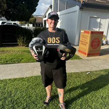 Wicked Boxing - A woman in casual athletic clothing holds two boxing helmets outdoors, with a building and greenery in the background. - Sporting Goods Near Me - Oxnard, California
