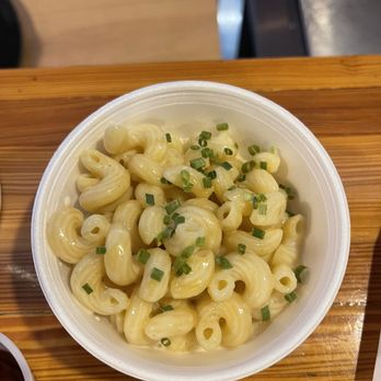 Kula's It's Just Sauce - Mac n cheese - A white disposable cup contains macaroni and cheese with elbow pasta coated in cheese sauce, garnished with chopped chives. The cup is positioned on a wooden surface, likely in a casual dining setting, with part of another cup visible at the bottom edge of the image. - Barbeque Near Me - Walker, Iowa
