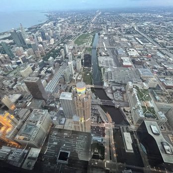 Skydeck Chicago - A high-altitude photograph captures the Chicago skyline, including numerous skyscrapers, the Chicago River flowing through the city, and the surrounding urban landscape extending toward Lake Michigan in the distance. The image shows a mix of modern and older buildings, with streets and bridges visible below, taken from a tall building or observation deck. - Observatories Near Me - Chicago, Illinois