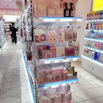 Shibuyala - A retail display shelf in a store features Sakura-themed skincare and beauty products, including boxed items, bottles, and plush accessories. The background has a pink floral design with the word 'SAKURA' prominently displayed. The shelf is illuminated with blue lighting and contains various beauty products, such as facial masks, creams, and sprays, arranged neatly. The store has a bright, clean interior with marble flooring, and other shelves with products are visible in the background. - Cosmetics & Beauty Supply Near Me - Boston, Massachusetts