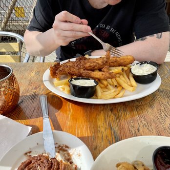 River Falls Restaurant - Fish & Chips - A person is eating fish and chips with fries and dipping sauces at an outdoor dining area, with a wooden table and metal chairs. - Italian Near Me - Woonsocket, Rhode Island