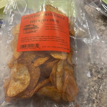 Delicious Beef Jerky - The image shows a clear plastic bag filled with potato chips seasoned with red chile powder. The bag has an orange label with the text "Delicious Red Chile Potato Chips" and includes ingredients such as potatoes, red chile powder, salt, pepper, and other seasonings. The label also provides the processing address in Albuquerque, NM, and the net weight of 4 ounces (113.39 grams). The chips are visible through the bag, and the background features a textured surface, possibly a countertop. - Meat Shops Near Me - Albuquerque, New Mexico