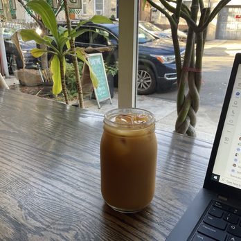 Daytime - Cold Brew - A mason jar filled with iced coffee and ice cubes is placed on a wooden table inside a cafe. The background shows large windows with plants and a view of parked cars and buildings outside. Part of a laptop is visible on the right side of the image, indicating a workspace or casual setting. - Sandwiches Near Me - Brooklyn, New York