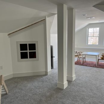 Dream House Services - Indoor Playhouse - A bright attic room features white walls, a sloped ceiling, and large windows allowing natural light to fill the space. A small white table with four chairs is positioned on a reddish rug near the windows. The room has gray carpet flooring and minimal furnishings, with a small window on the left wall and structural columns dividing the space. - General Contractors Near Me - Randolph, Massachusetts