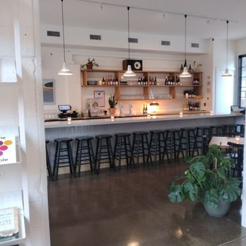 Out There - The image shows a bar interior with a white countertop and a row of black stools. Behind the bar, wooden shelves display bottles of alcohol, glasses, and small potted plants. Pendant lights hang from the ceiling, providing lighting. The space has a modern, minimalist design with a polished concrete floor and a ceiling fan. A large window on the right allows natural light into the room. - Wine Bars Near Me - Sawyer, Michigan