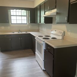 Before - A kitchen features dark-colored cabinets, a window above the sink, and white appliances such as a stove and range hood, with light-colored countertops and flooring.