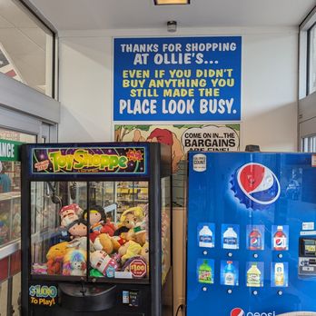 Ollie's Bargain Outlet - The image shows the interior of Ollie's Bargain Outlet, featuring a claw machine filled with plush toys and a Pepsi vending machine. The claw machine is labeled with a $1.00 to play sign, and the vending machine displays various drinks. There are signs on the wall, including a blue sign with shopping humor and a partial view of the store entrance with glass doors and windows. - Discount Store Near Me - Barkhamsted, Connecticut